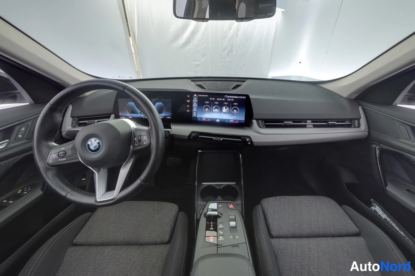 Etuistuimet: Cabin view of a car with a black dashboard featuring a dual-screen setup, a multi-function steering wheel with buttons, and fabric front seats. Center console i