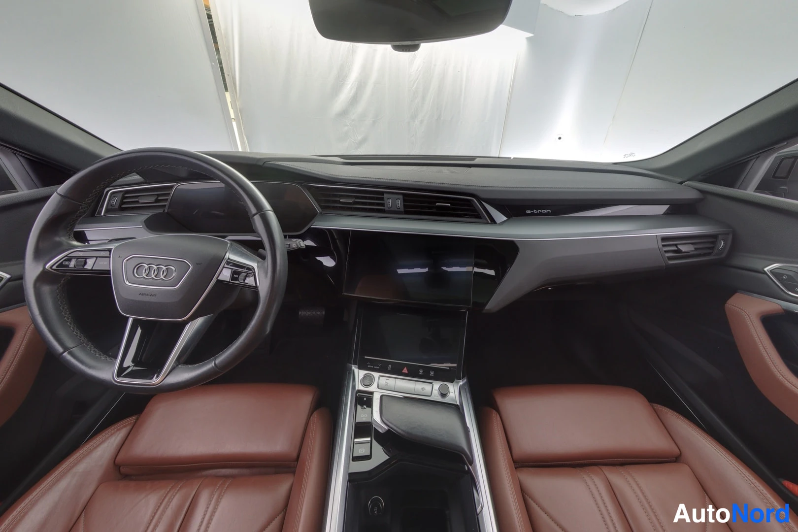 Ohjaamo: Kuvassa näkyy automobiilin ohjaamo: ohjauspyörä (Audi-logo), mittaristo, keskimmäinen kosketusnäyttö ja nahkapäällysteiset istuimet (ruskeat).