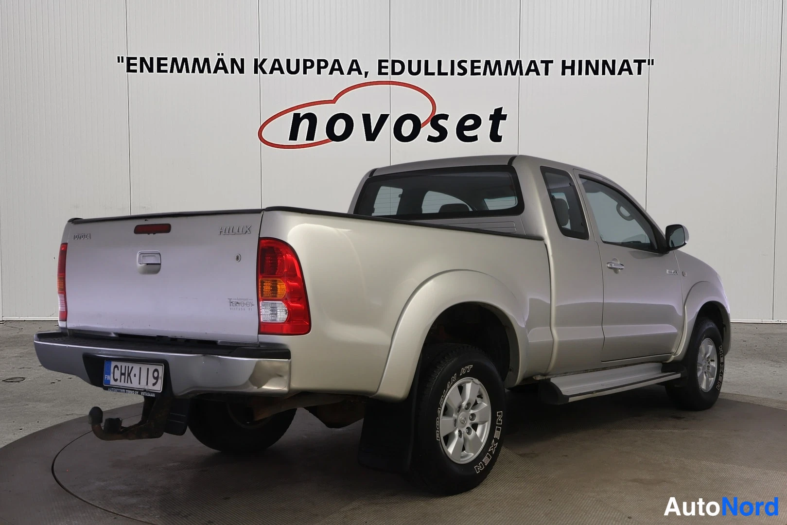 Takaviisto: Hopeanvärinen Toyota Hilux kuvattuna takaviistosta; lava, takavalot ja rekisterilauta näkyvät, kylki kuvan oikealla puolella.