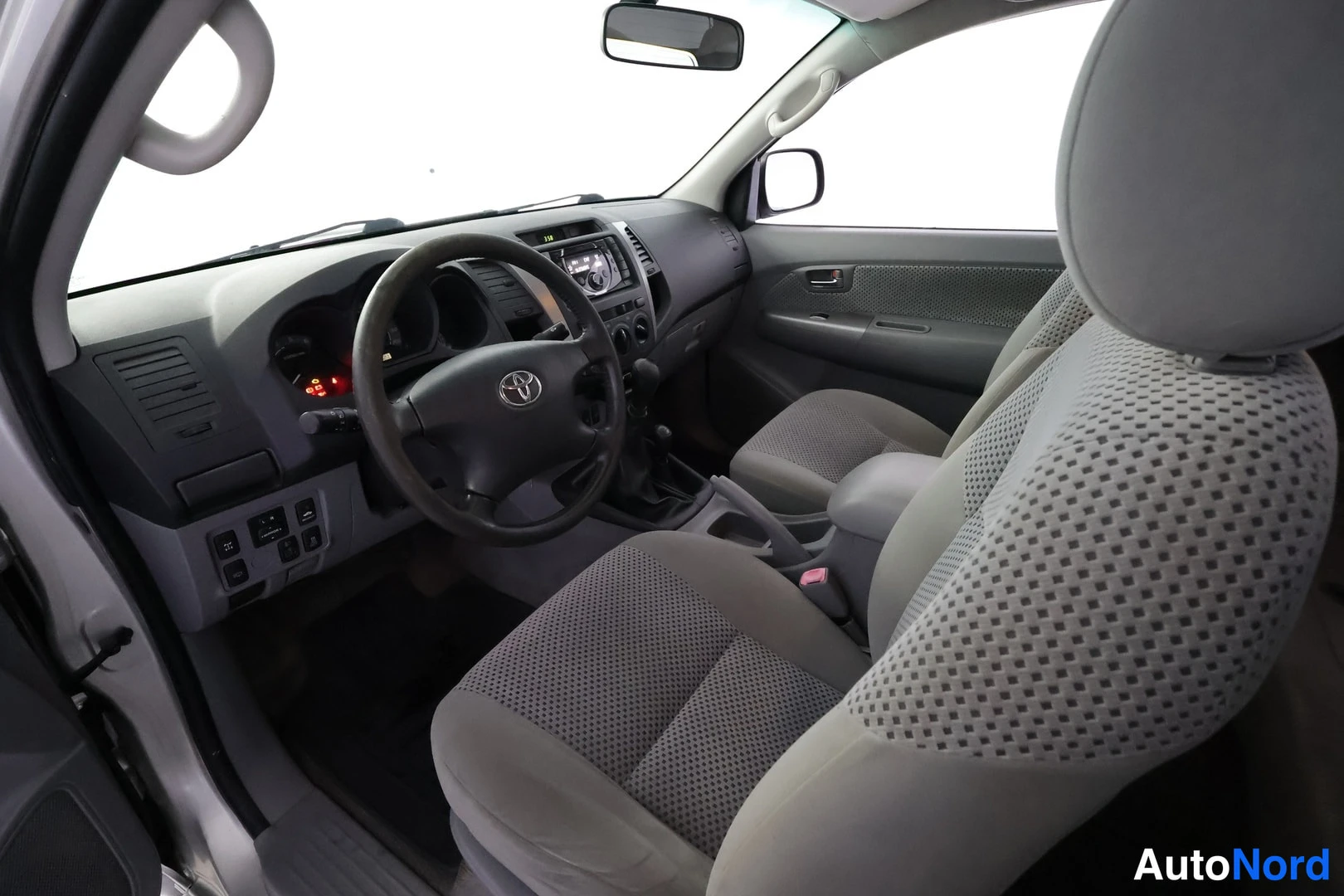 Etuistuimet: Ohjaamosta kulmasta kuvattuna: ohjauspyörä Toyota-merkillä, kojelauta, keskusyksikkö radioineen ja harmaat kangasistuimet.