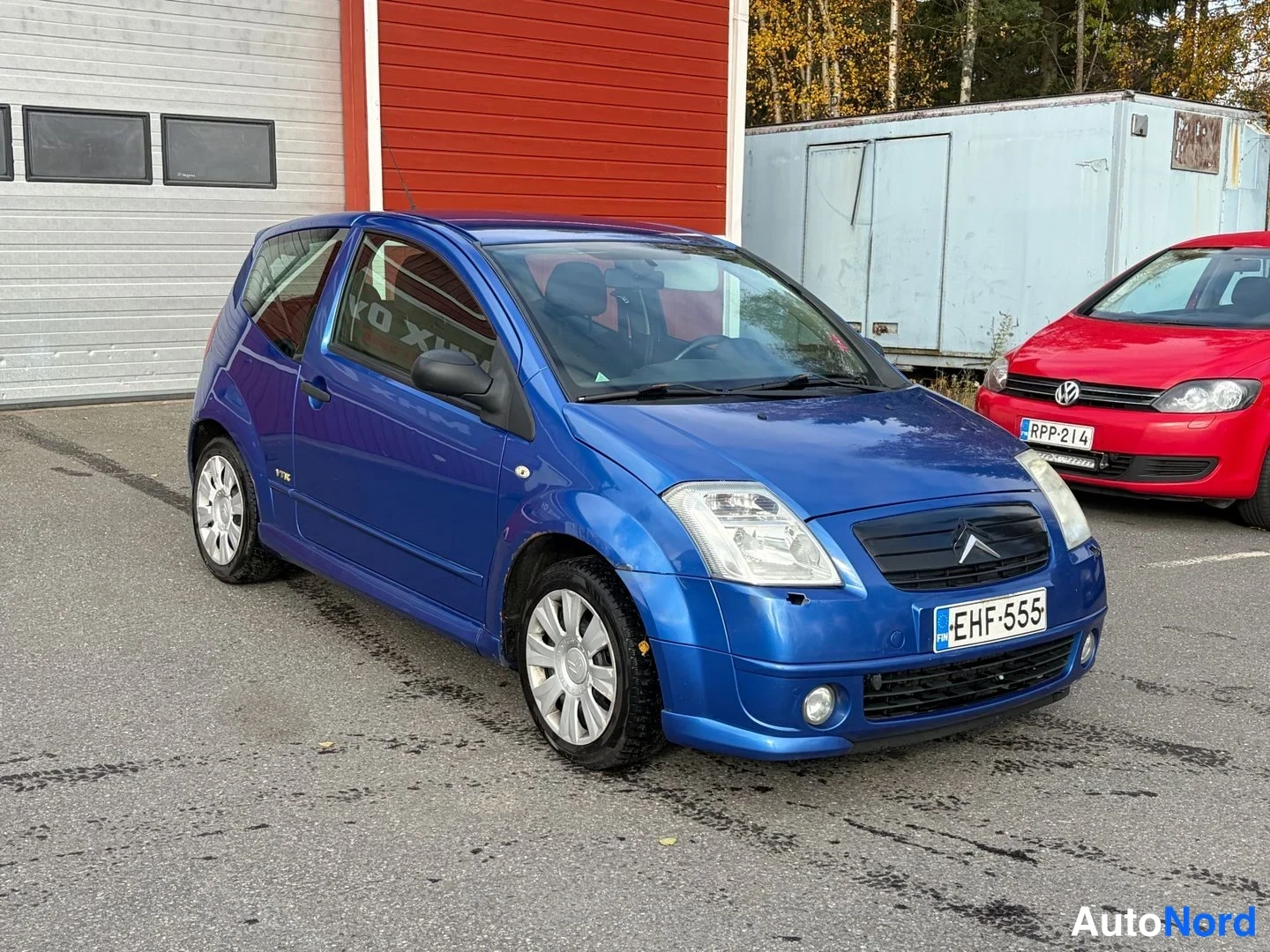 Etuviisto, nokka oikealle: Sininen Citroën C2 etukulmakuvassa oikealta päin kuvattuna, rekisteritunnus, taustalla punainen henkilöauto.