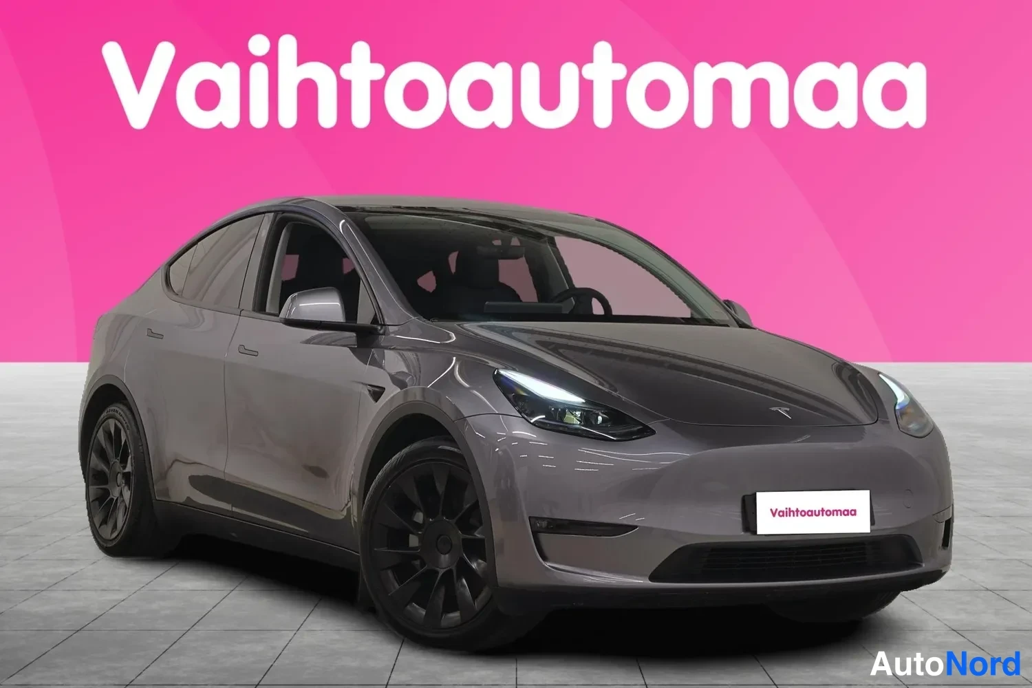 Etuviisto, nokka oikealle: Tesla Model Y etukulmasta oikealta, tummanharmaa koriväri, mustat java-tyyppiset peltivanteet ja LED-ajovalot päällä.