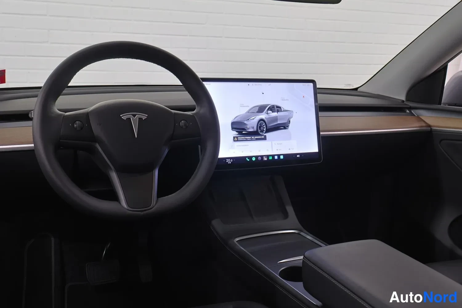 Edestä: Tesla-merkkinen ohjauspyörä ja keskellä kosketusnäyttö, jossa näkyy ajoneuvon kuva ja valikko; tumma sisusta ja puunvyöhyke kojelaudassa.