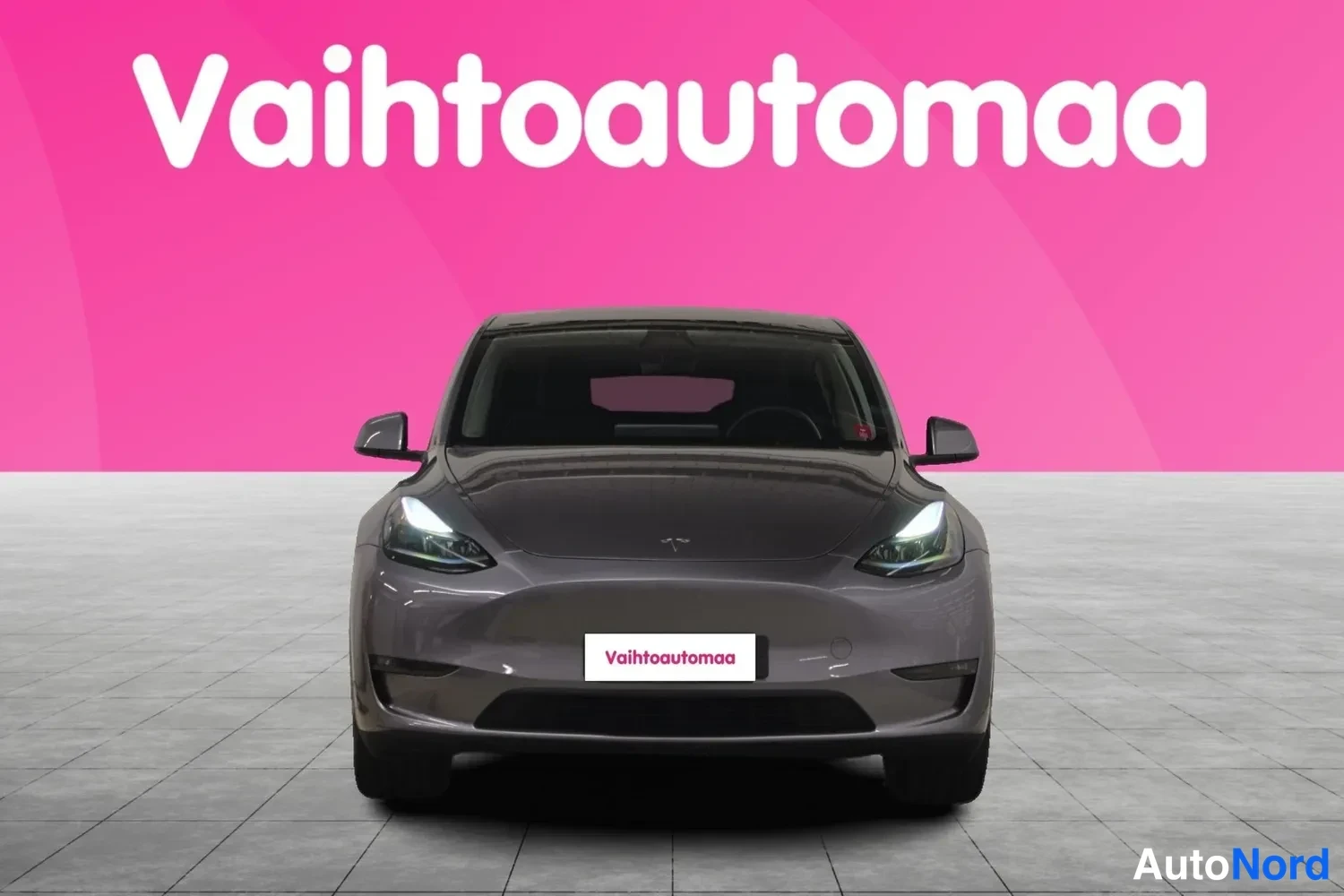 Edestä: Tesla Model Y suoraan edestä kuvattuna, tummanharmaa koriväri, LED-ajovalot päällä ja Vaihtoautomaan kilpi etupuskurissa.