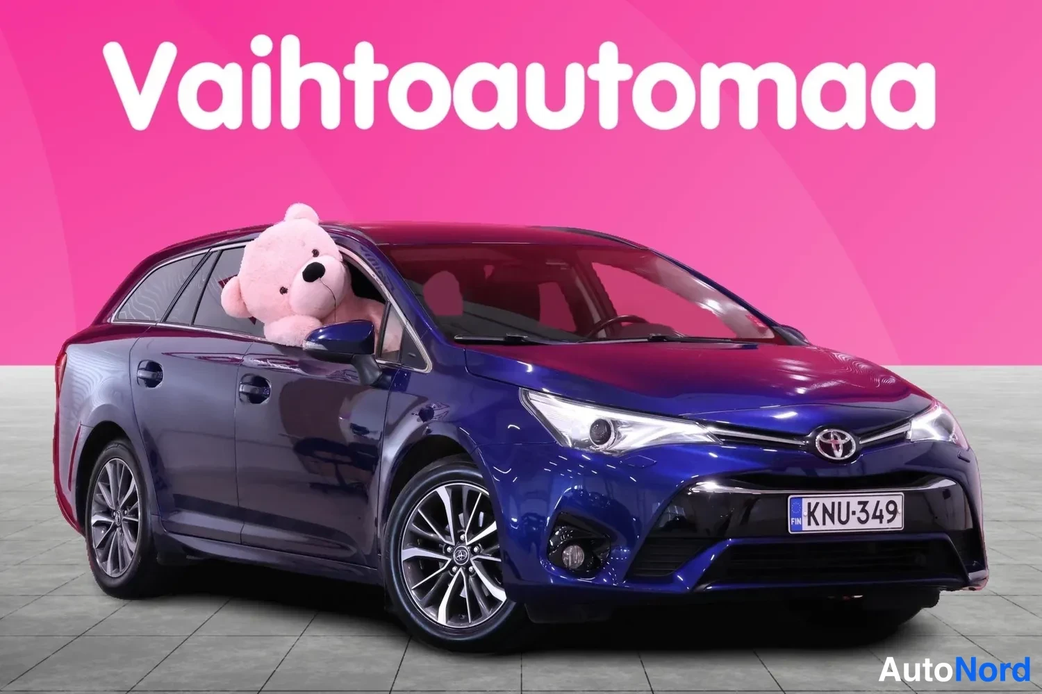 Etuviisto, nokka oikealle: Tummansininen Toyota Avensis -farmari etuviistosta, keula oikealla; Toyota-merkki ja rekisterikilpi näkyvät keulalla ja vaaleanpunainen pehmokarhu kurkis