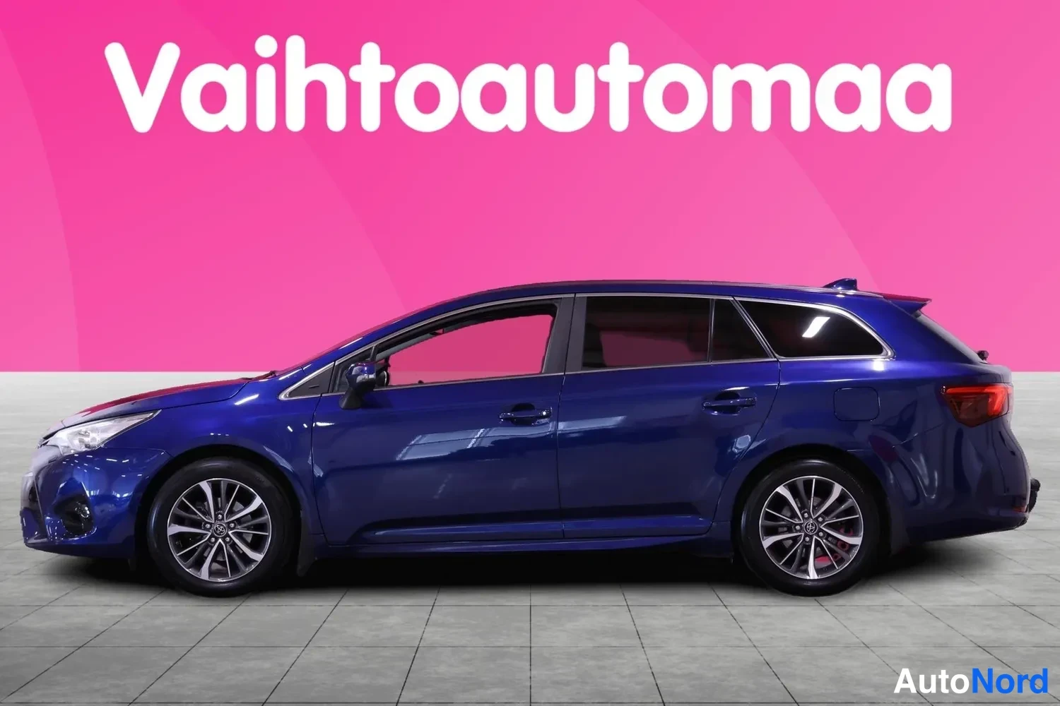 Etuviisto, nokka vasemmalle: Sininen Toyota Avensis -farmari sivulta vasemmasta kulmasta kuvattuna, kevytmetallivanteet näkyvissä.