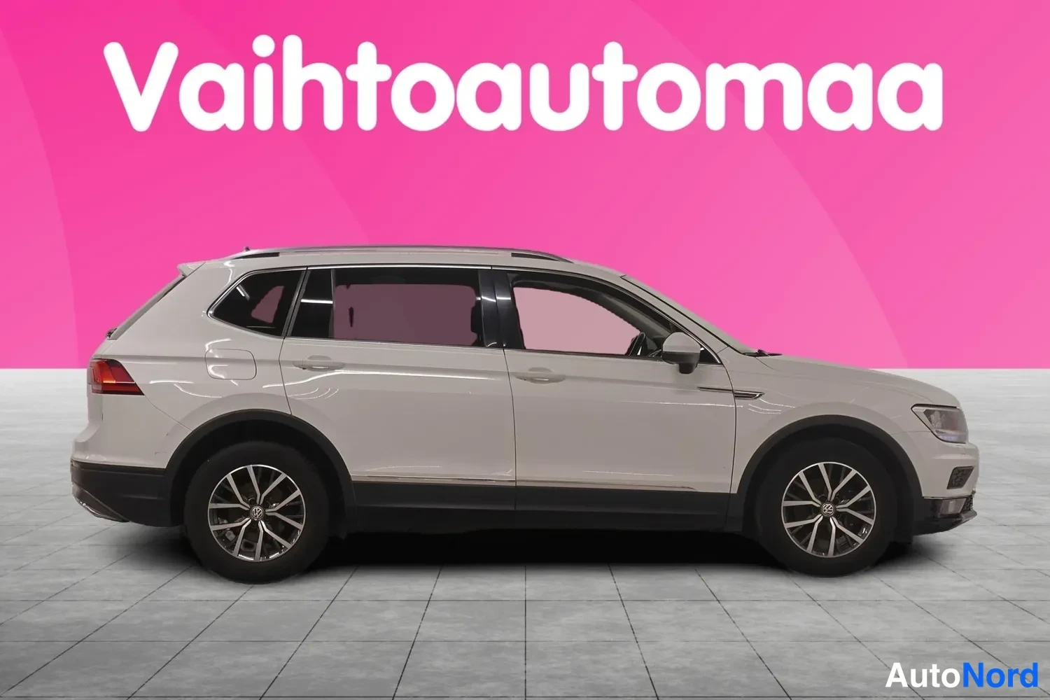 Etuviisto, nokka oikealle: Valkoinen Volkswagen Tiguan Allspace sivulta kuvattuna, oikean puolen ovet ja pyörät näkyvissä, vaaleanpunainen tausta.