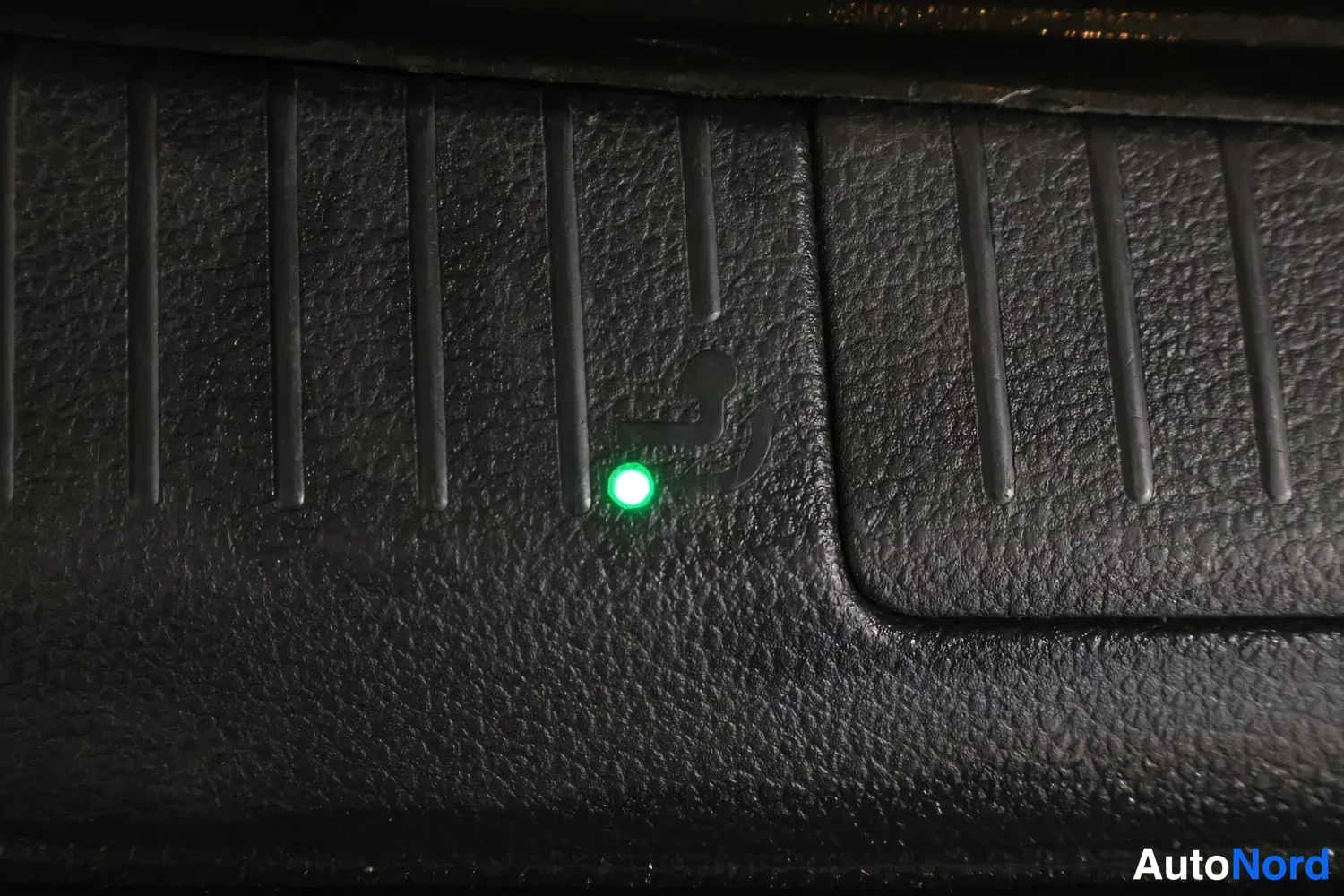 Yksityiskohta: Close-up view of a black textured dashboard panel showing an illuminated green indicator light next to an embossed seatbelt warning symbol.