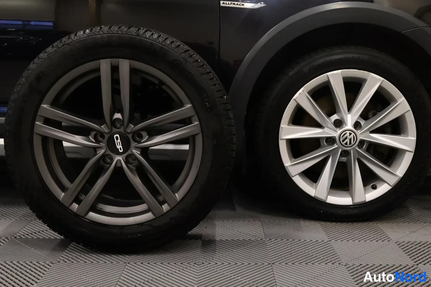 Sivulta: A dark grey vehicle's side wheel arch and wheel are visible. Beside it, a spare tyre with matching rims stands vertically on a tiled floor.