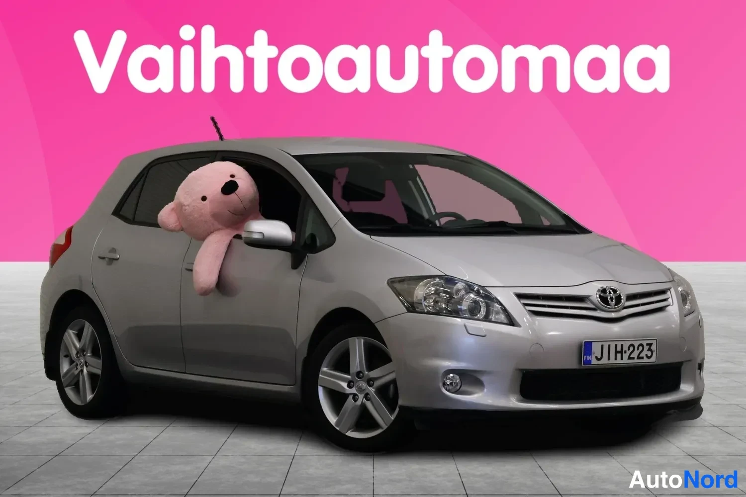 Etuviisto, nokka vasemmalle: Hopeanvärinen Toyota Auris etuosaltaan ja sivuiltaan näkyvissä, rekisteritunnus, vaaleanpunainen tausta.