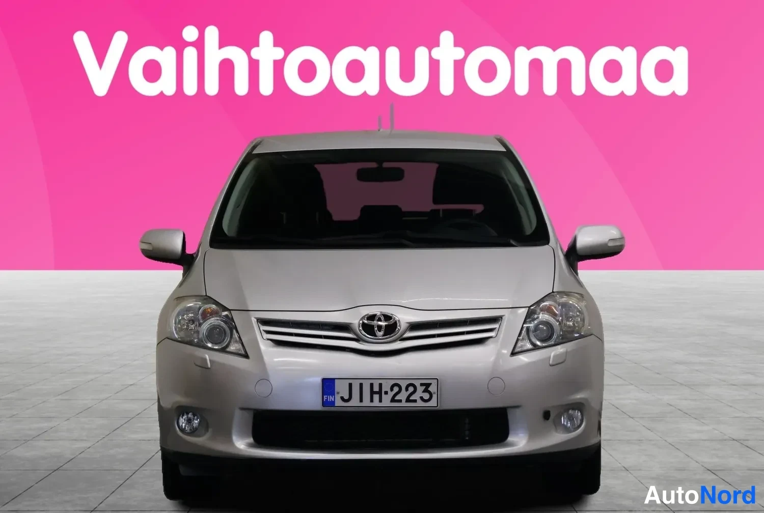 Edestä: Hopeanvärinen Toyota Auris suoraan edestä, rekisteritunnus, vaaleanpunainen tausta.