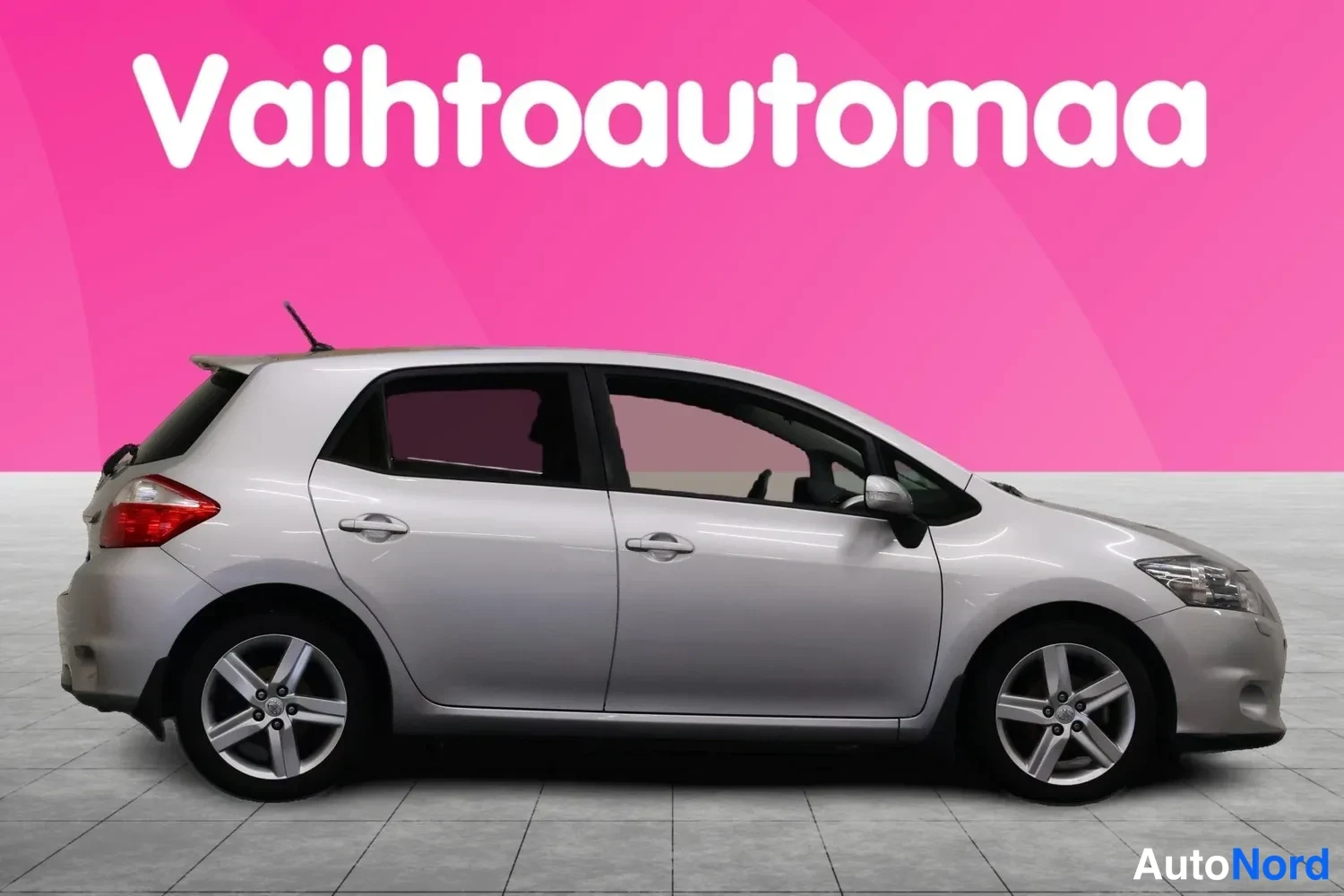 Sivulta: Hopeanharmaa viistoperäinen henkilöauto sivusta kuvattuna, radioasema Vahtoautomaan mainosbannerin edessä.