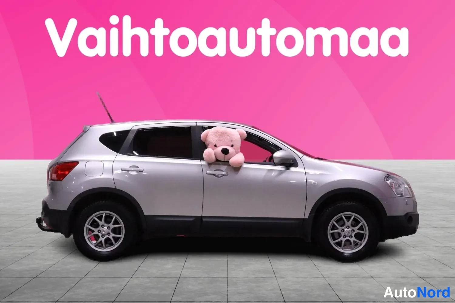 Sivulta: Hopeanvärinen Nissan Qashqai sivulta vasemmasta kyljestä kuvattuna studiossa, taustalla vaaleanpunainen mainoskangas tekstillä Vaihtoautomaa.