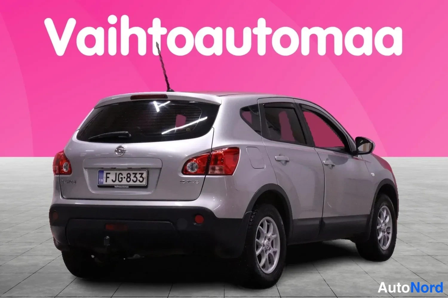 Takaviisto: Hopeanvärinen Nissan Qashqai takaa viistosti vasemmalta näkyvässä studiossa; rekisterikilpi, taustalla vaaleanpunainen mainoskangas tekstillä Vaihtoautomaa.