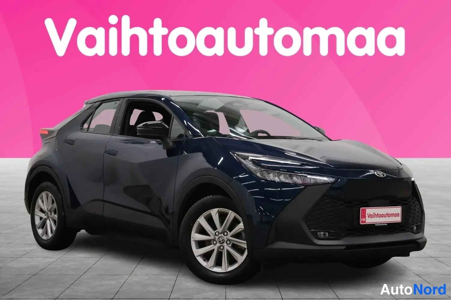 Etuviisto, nokka oikealle: Sininen Toyota C-HR etukulmasta oikealta, myyjän tunnisteet ja pinkki tausta.