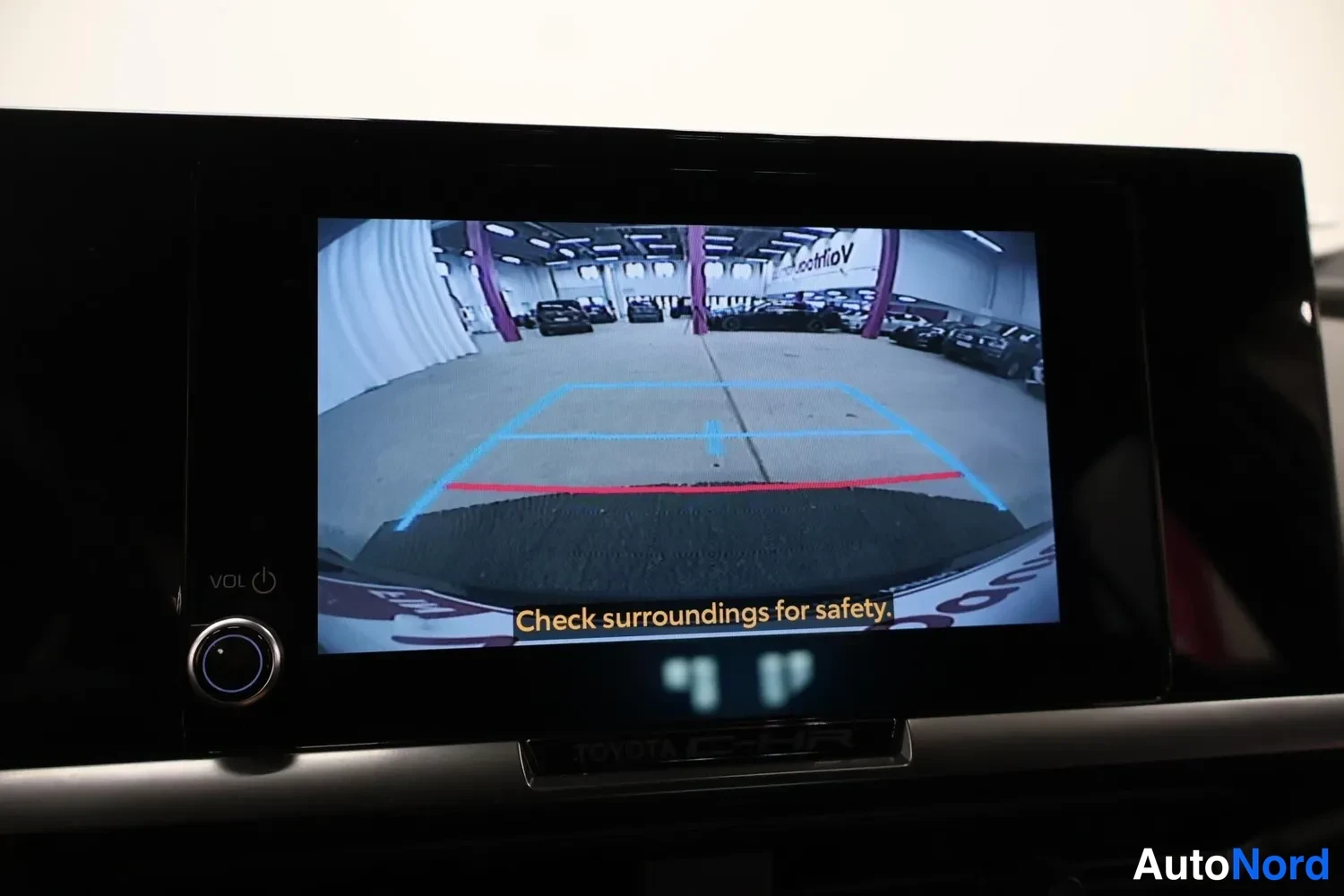 Yksityiskohta: Infotainment-näyttö, jossa näkyy peruutuskameran kuva ohjausviivoineen ja tekstillä Check surroundings for safety.