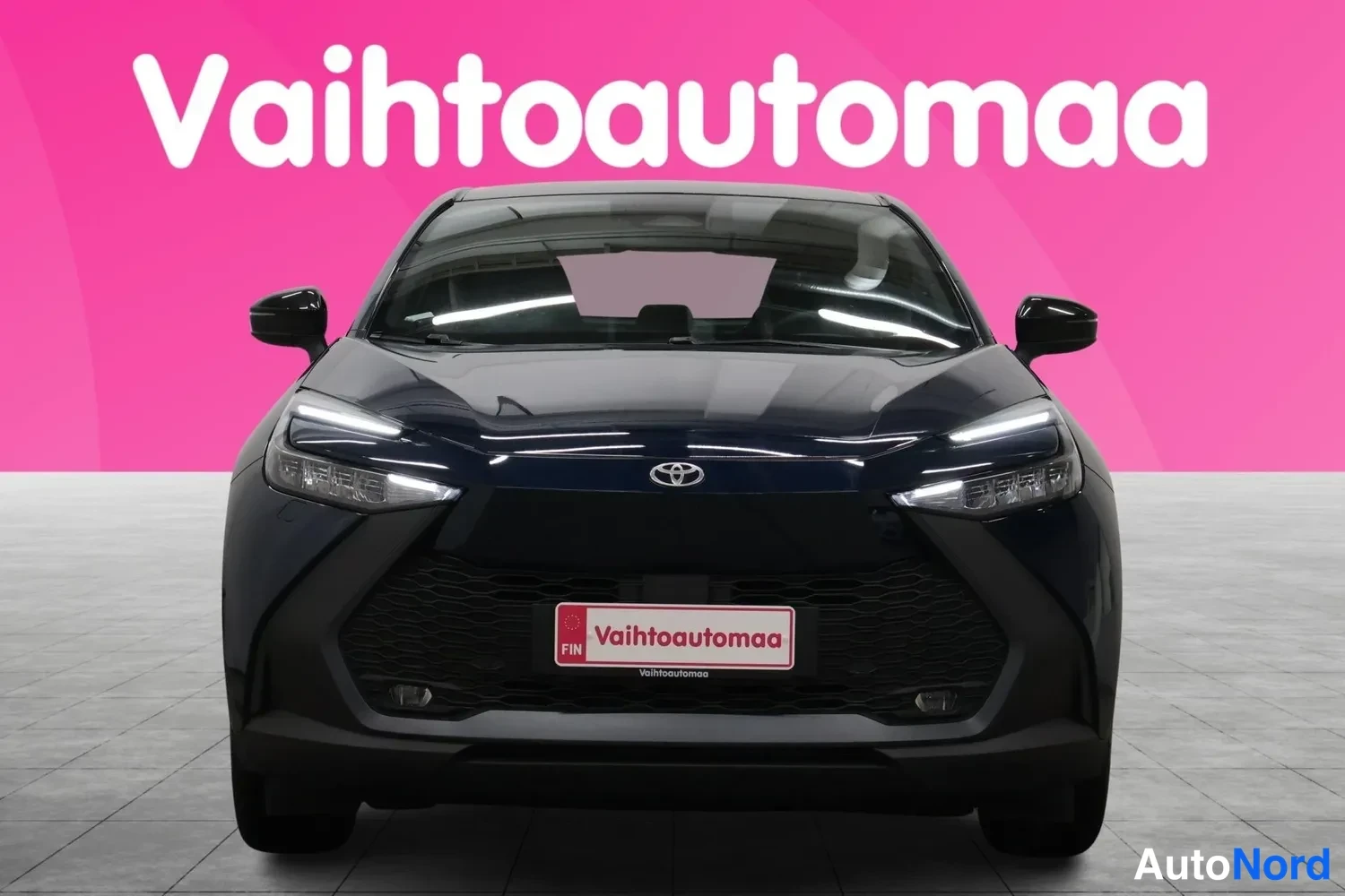 Edestä: Tummansininen Toyota C-HR suoraan edestäpäin, pinkki tausta ja Vaihtoautomaan tunnisteet.
