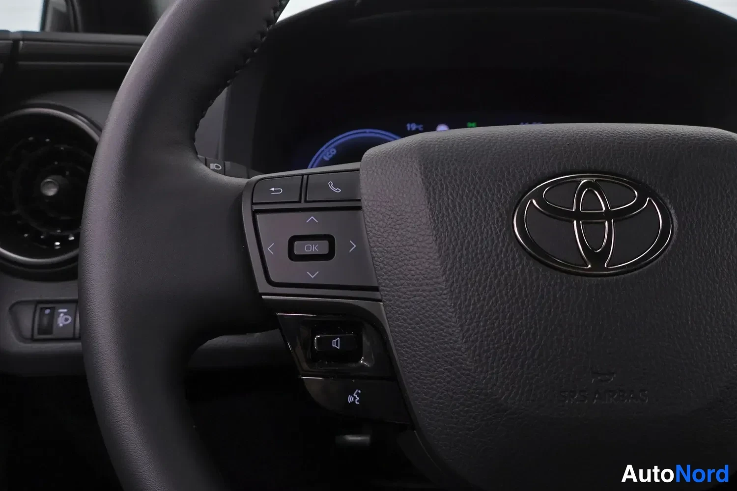 Yksityiskohta: Lähikuva ohjauspyörästä, Toyota-merkki ja SRS AIRBAG -teksti näkyvät, vasemmalla pikanäppäimet.