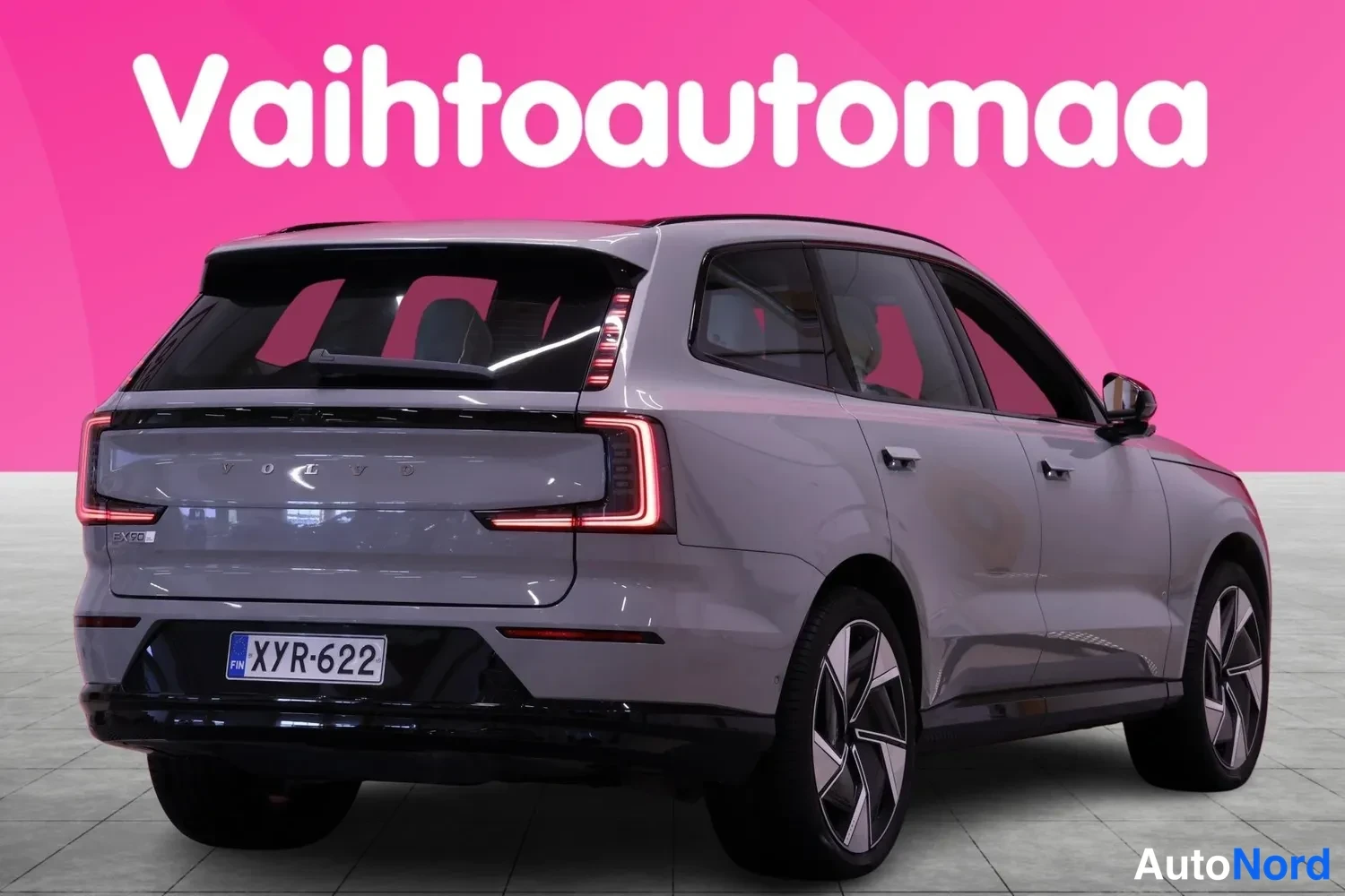 Takaviisto: Auton takakulma: vaaleanharmaan tila-autoon takaosa ja vasen kylki, rekisterikilpi, vaaleanpunainen tausta.