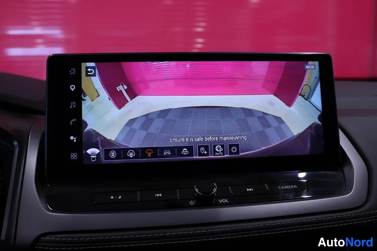 Yksityiskohta: Infotainment-näytöllä näkyy takakameran laajanakulmakuva punaisessa tilassa, teksti 'Ensure it is safe before manoeuvring'.