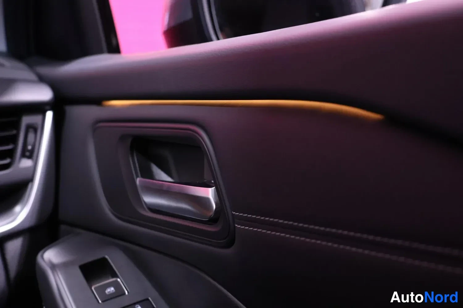 Ohjaamo: Close-up view of a car's interior door panel showing an integrated door handle with metallic finish, power window switch with two buttons, and illuminated ambie