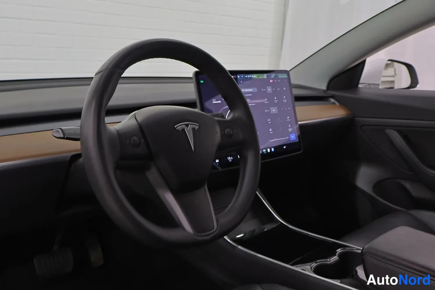 Muu: Tesla Model 3:n ohjaamo: ratin takana keskellä ohjauspaneeli ja suuri keskikonsolin kosketusnäyttö, puinen koristelista kojelaudassa.