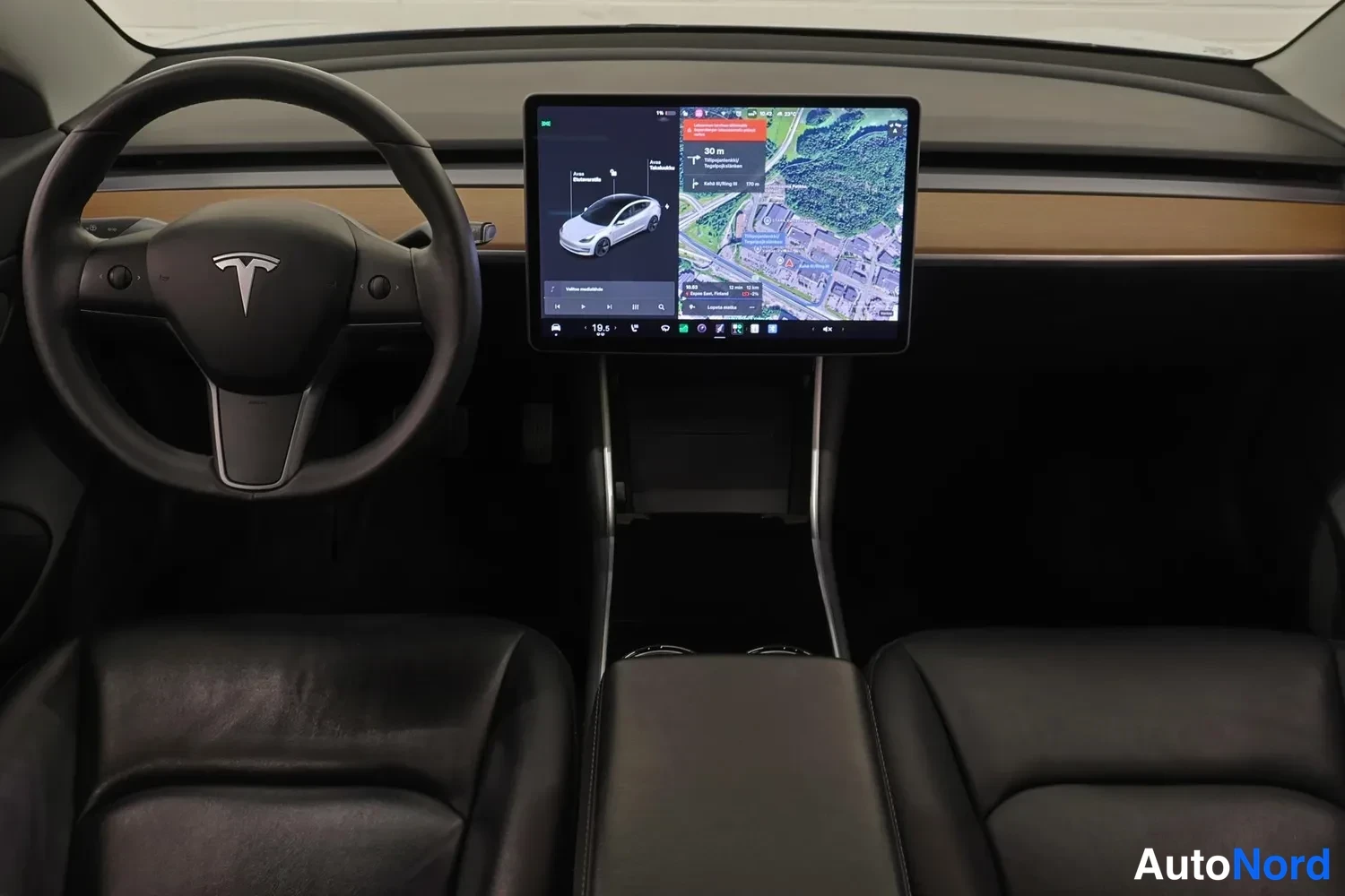 Muu: Tesla Model 3:n kojelauta suoraan edestä: ratti vasemmalla, keskellä kosketusnäyttö, jossa näkyy kartta ja autossa tehtäviä asetuksia; puinen koristelista ja mu