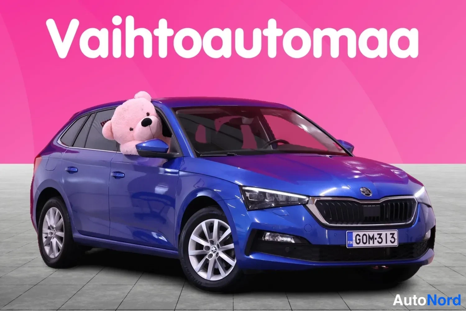 Etuviisto, nokka oikealle: Sininen Skoda Scala esitettynä etukulmasta oikealle suunnattuna, pinkki Vahtoautomaa-tausta ja pehmolelu kuljettajan ikkunassa.