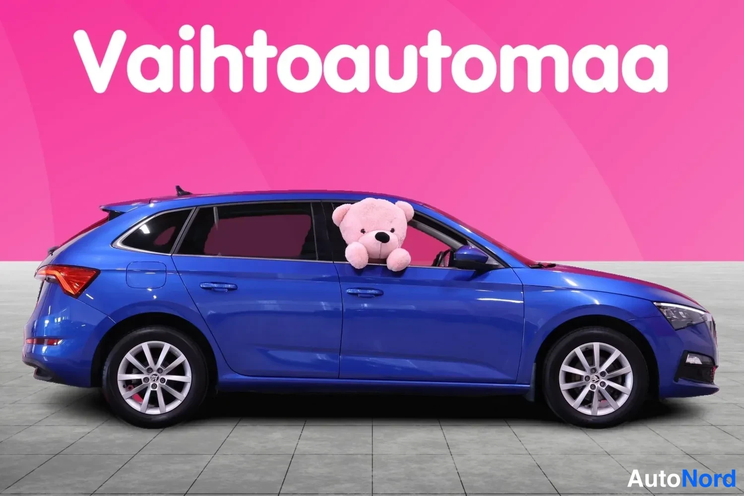 Sivulta: Sininen Skoda Scala sivuprofiilista vasemmalta kuvattuna, pehmolelu takamatkustajan ikkunassa, pinkki tausta.
