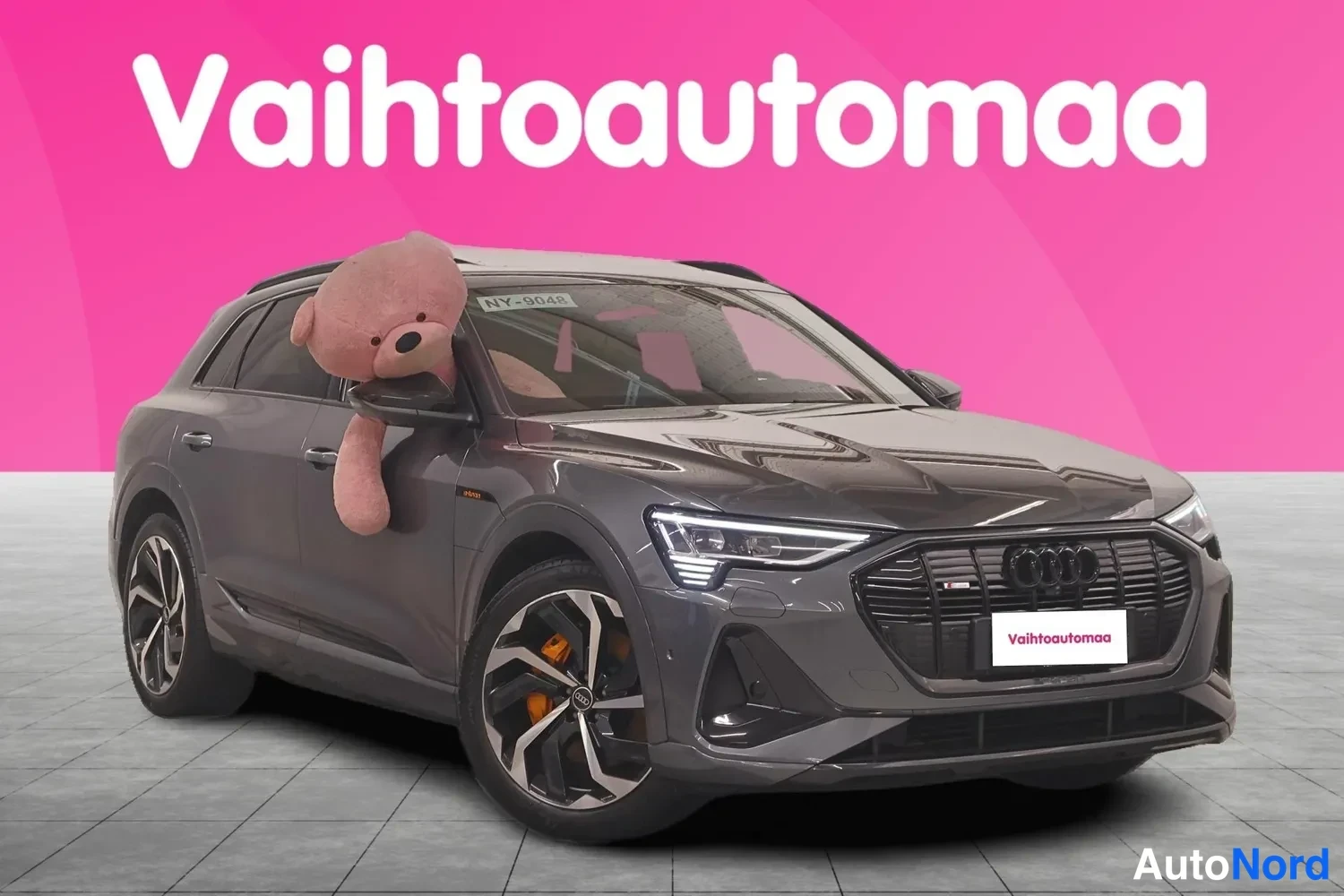 Etuviisto, nokka oikealle: Tummanharmaa Audi e-tron -maasturikuva etukulmasta oikealta, etusäleikkö ja kylki näkyvissä, pinkki tausta.