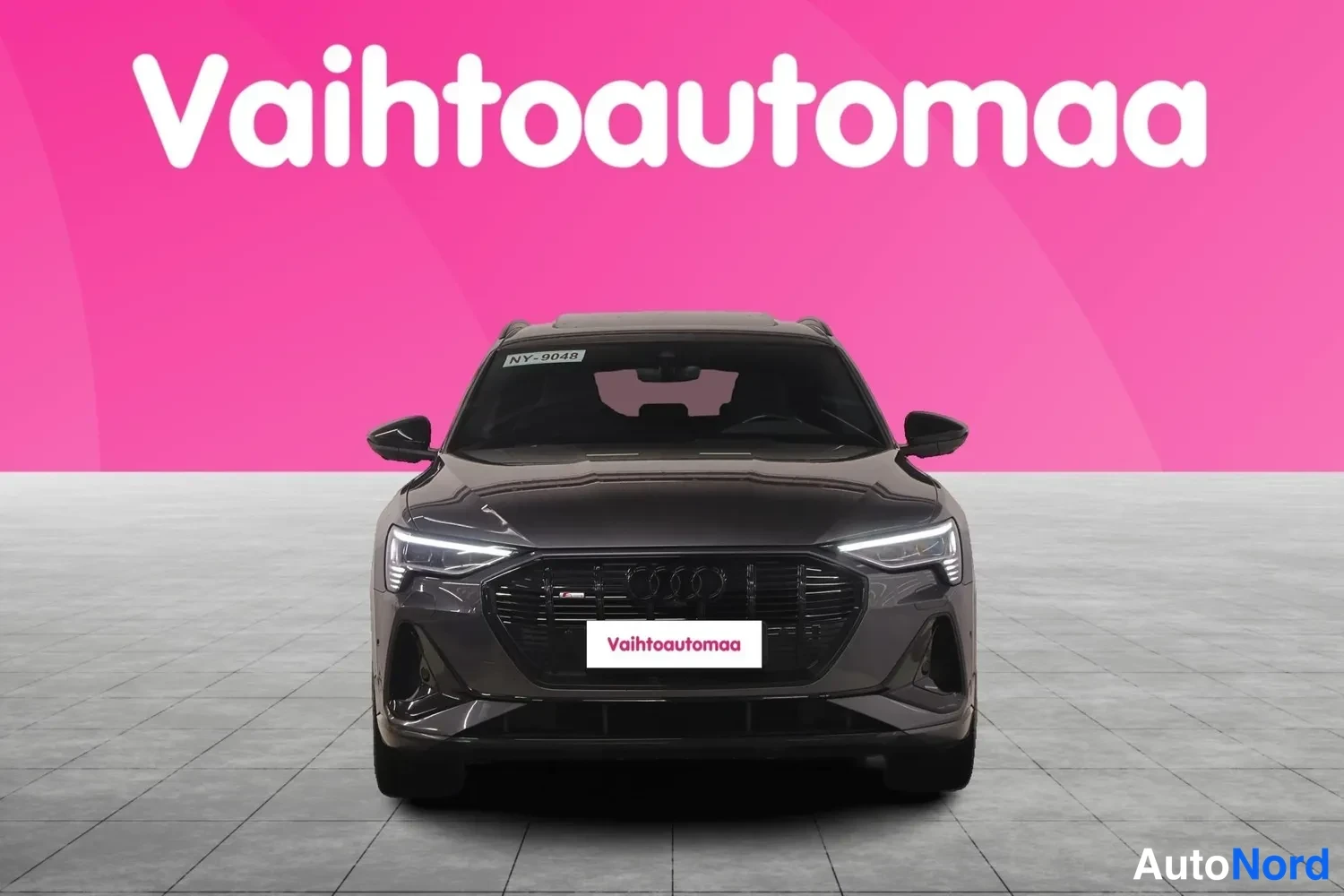 Edestä: Tummanharmaan Audi e-tronin suora etukuva, etusäleikkö ja LED-ajovalot näkyvissä, pinkki tausta.