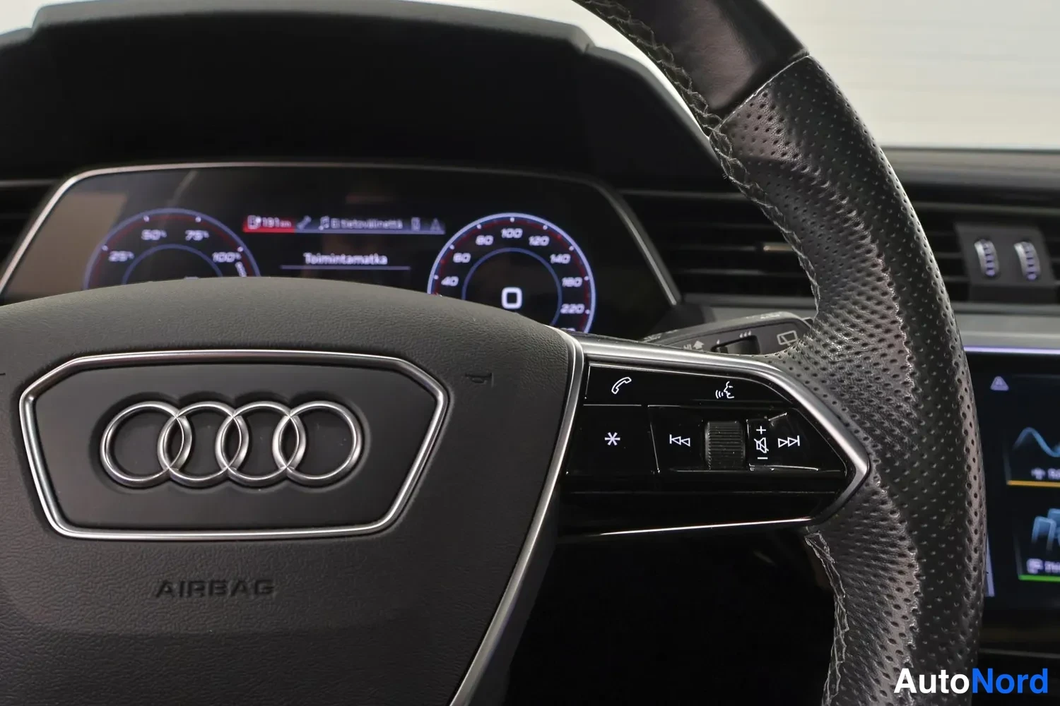Yksityiskohta: Audi-ohjauspyörä etualalla, taustalla digitaalinen mittaristo ja osa keskikonsolin näytöstä.