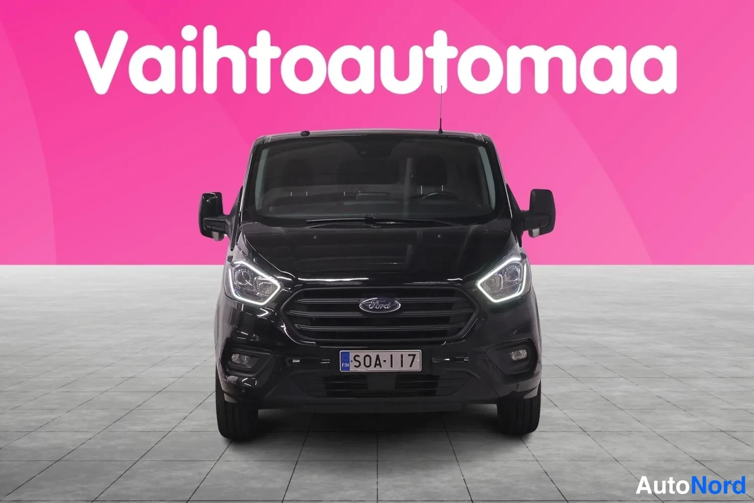 Edestä: Musta pakettiauto suoraan edestä valaistuilla ajovaloilla, rekisteritunnus.