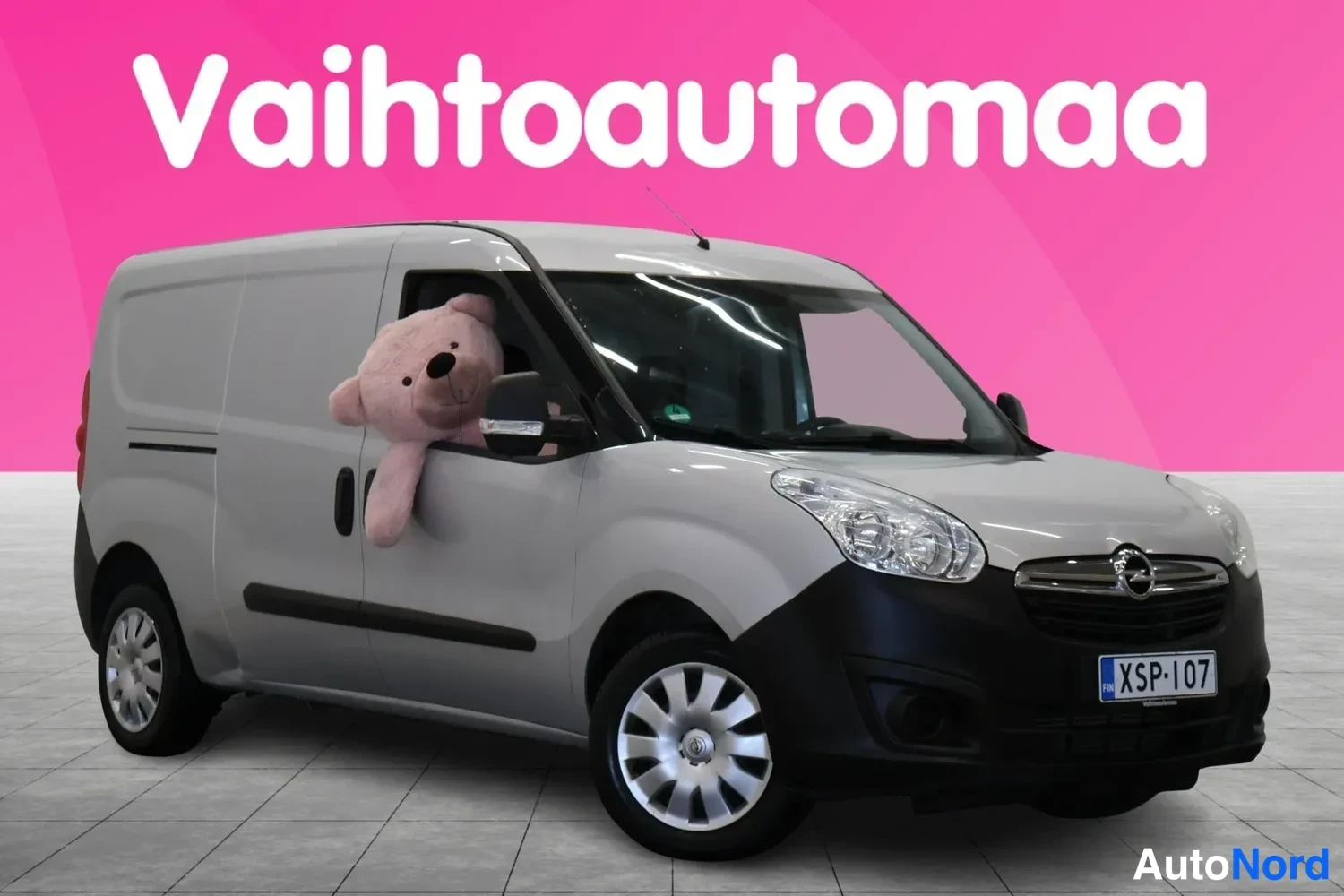 Etuviisto, nokka oikealle: Vaaleanharmaa pakettiauto etuviistosta oikealta, rekisterikilpi, taustana pinkki markkinointigrafiikka.