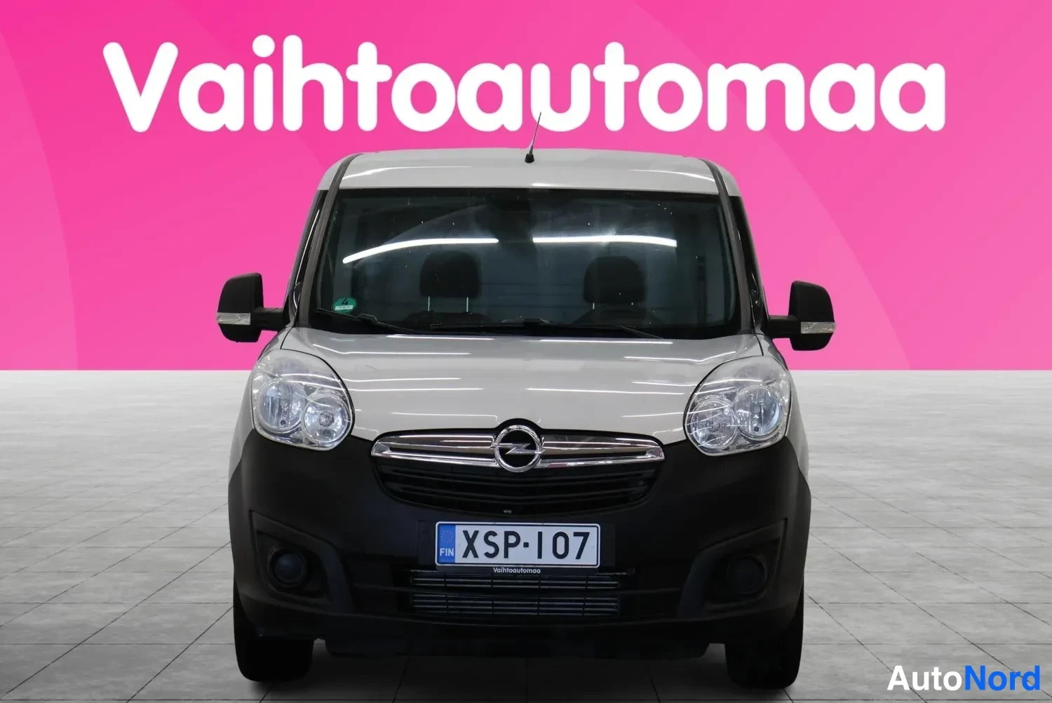 Edestä: Vaaleanharmaan pakettiauton etunäkymä, Opel-merkki ja rekisterikilpi, taustana pinkki markkinointigrafiikka.