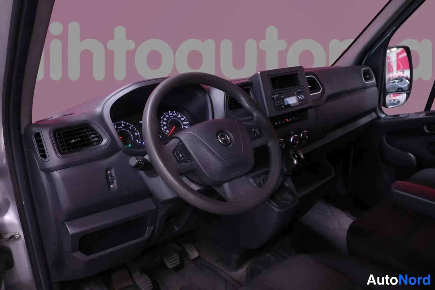 Muu: Avoimesta kuljettajan ovesta kuvattu pakettiauton sisätila: musta kojelauta, Opel-ohjauspyörä, mittarit ja radio.