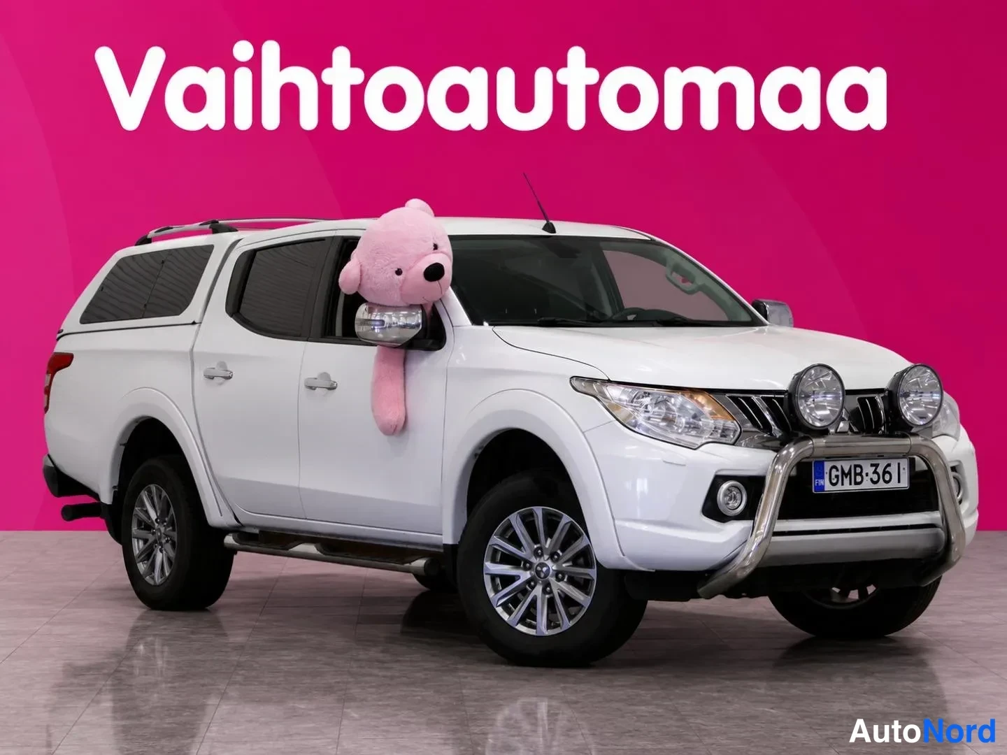 Etuviisto, nokka oikealle: Mitenkään valkoinen Mitsubishi L200 -avolava kuvattuna kolmanneskulmasta keula oikealla, pinkki tausta ja teksti Vaihtoautomaa.