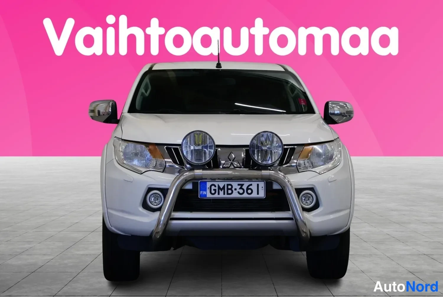 Edestä: Valkoisen Mitsubishi L200 -avolavan keula suoraan edestä, etupuskurissa kromikaari ja lisävalot, rekisteri, tausta pinkki.