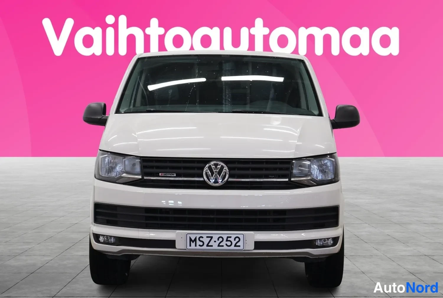 Edestä: Valkoisen Volkswagen Transporter -pakettiauton etunäkymä suoraan edestä; musta etusäleikkö ja Volkswagen-merkki, rekisteri.