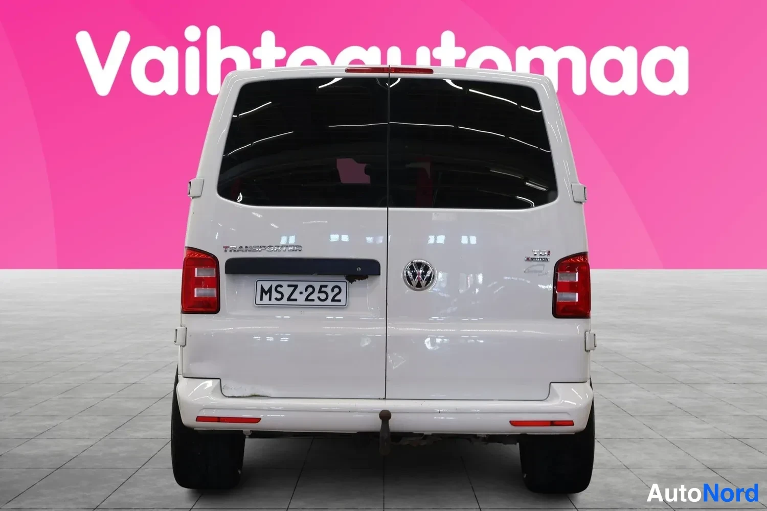 Takaa: Valkoinen pakettiauto suoraan takaa, rekisteritunnus, Volkswagen-merkki ja TRANSPORTER-teksti takaluukussa.