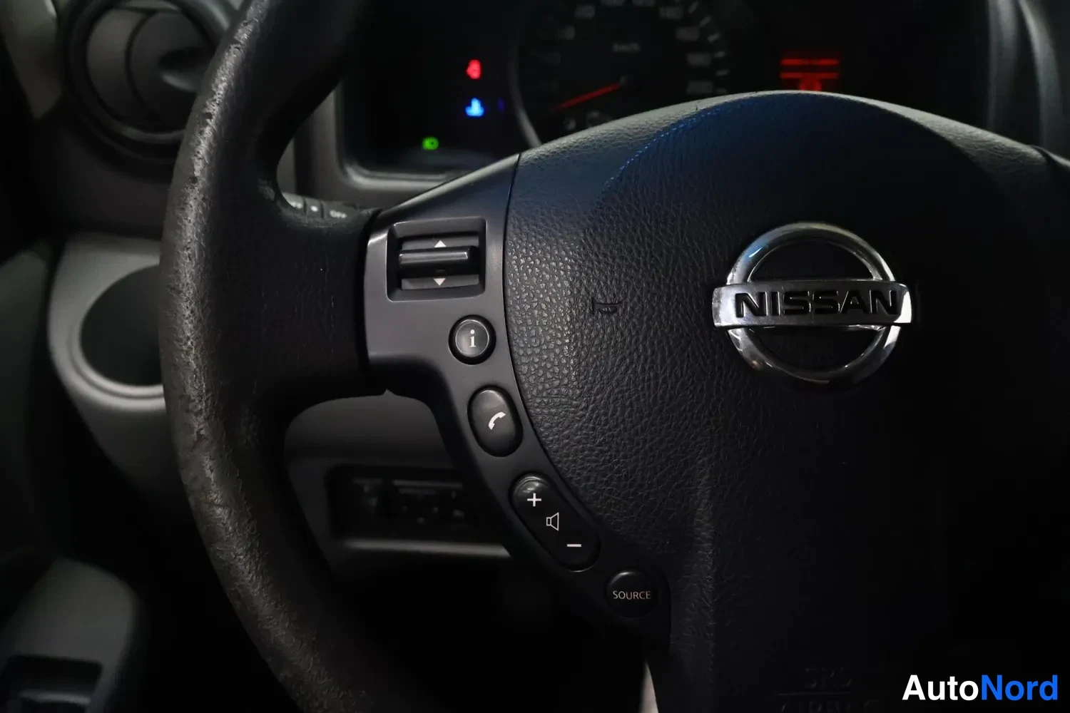 Muu: Nissan-merkkinen ohjauspyörä, jossa äänenvoimakkuuden ja puhelun nappeja ja SOURCE-merkitty painike.