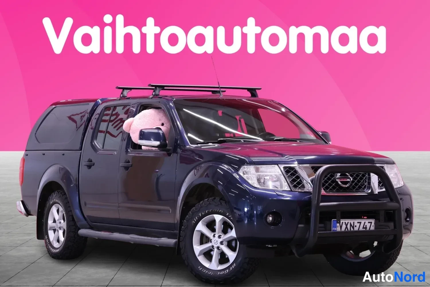 Etuviisto, nokka oikealle: Sininen Nissan Navara-noutoauto etukulmasta oikealta, kromikehys etupuskurissa ja kattoteline.