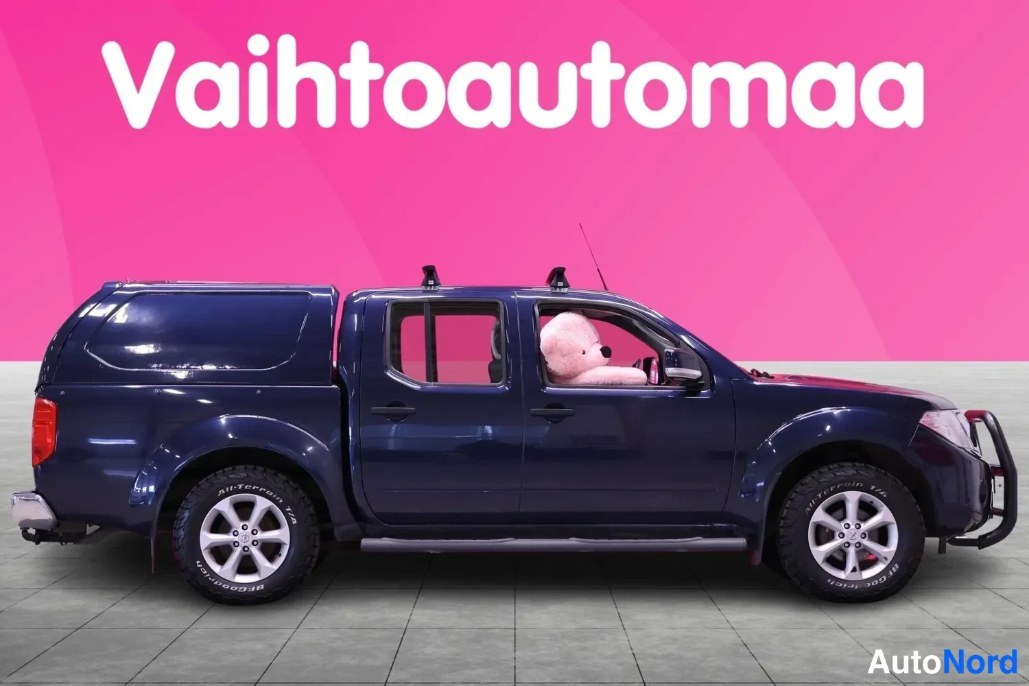 Etuviisto, nokka oikealle: Tummansininen Nissan Navara-noutoauto sivusta vasemmalta, katolla kattotelineet ja ohjaamossa pehmolelu.