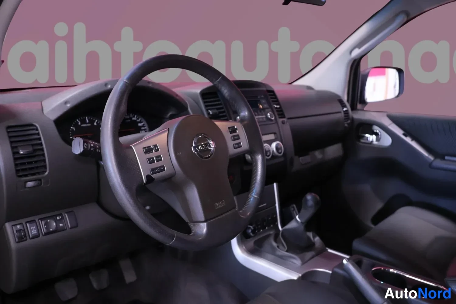 Muu: Auton sisätilan etunäkymä: Nissan-merkitty ohjauspyörä, mittaristo, manuaalivaihteen vaihdevipu, tummat sisäpaneelit ja keskikonsolin kaiutinjärjestelmä.