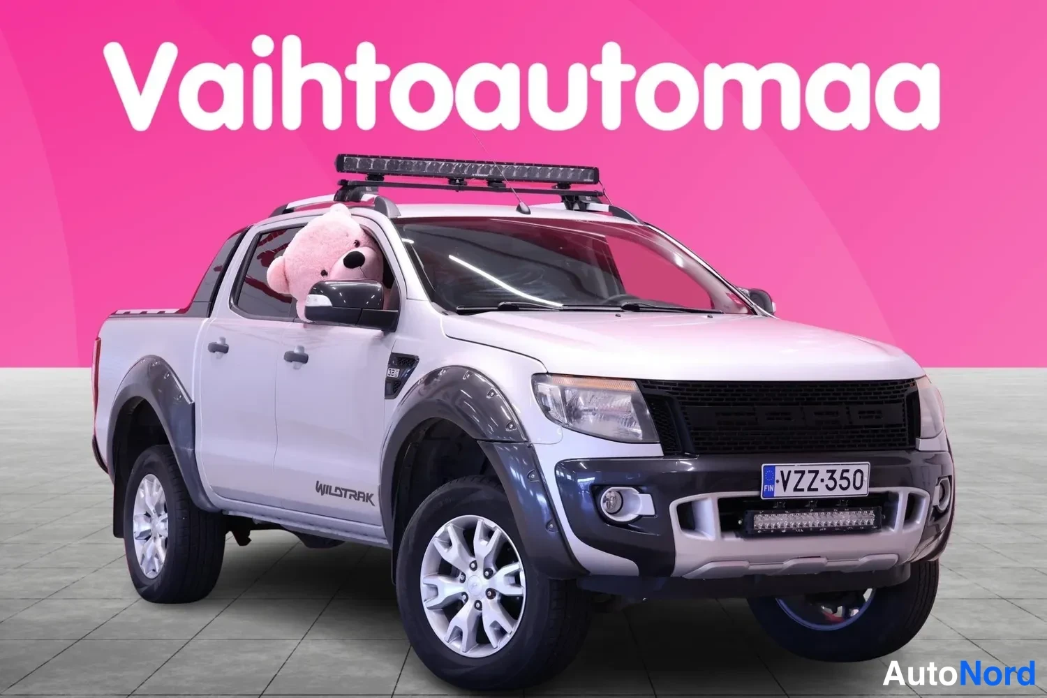 Etuviisto, nokka oikealle: Valkoinen parihaaraora Ford Ranger -mallinen avolava-auto kolmen neljäsosan etunäkymässä, kattokehillä ja keulaan asennetulla LED-valobarilla, taustalla