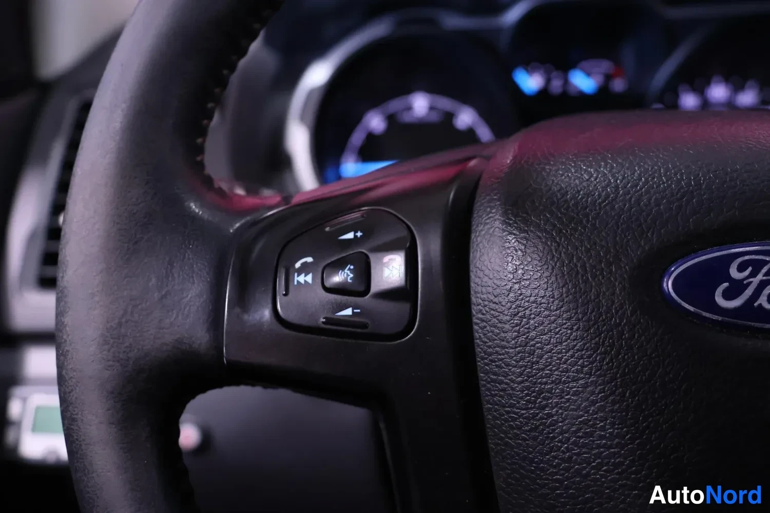 Muu: Ohjauspyörän vasemmanpuoleiset painikkeet puhelin- ja ääniohjaussymboleineen, Ford-emblem osittain näkyvää.
