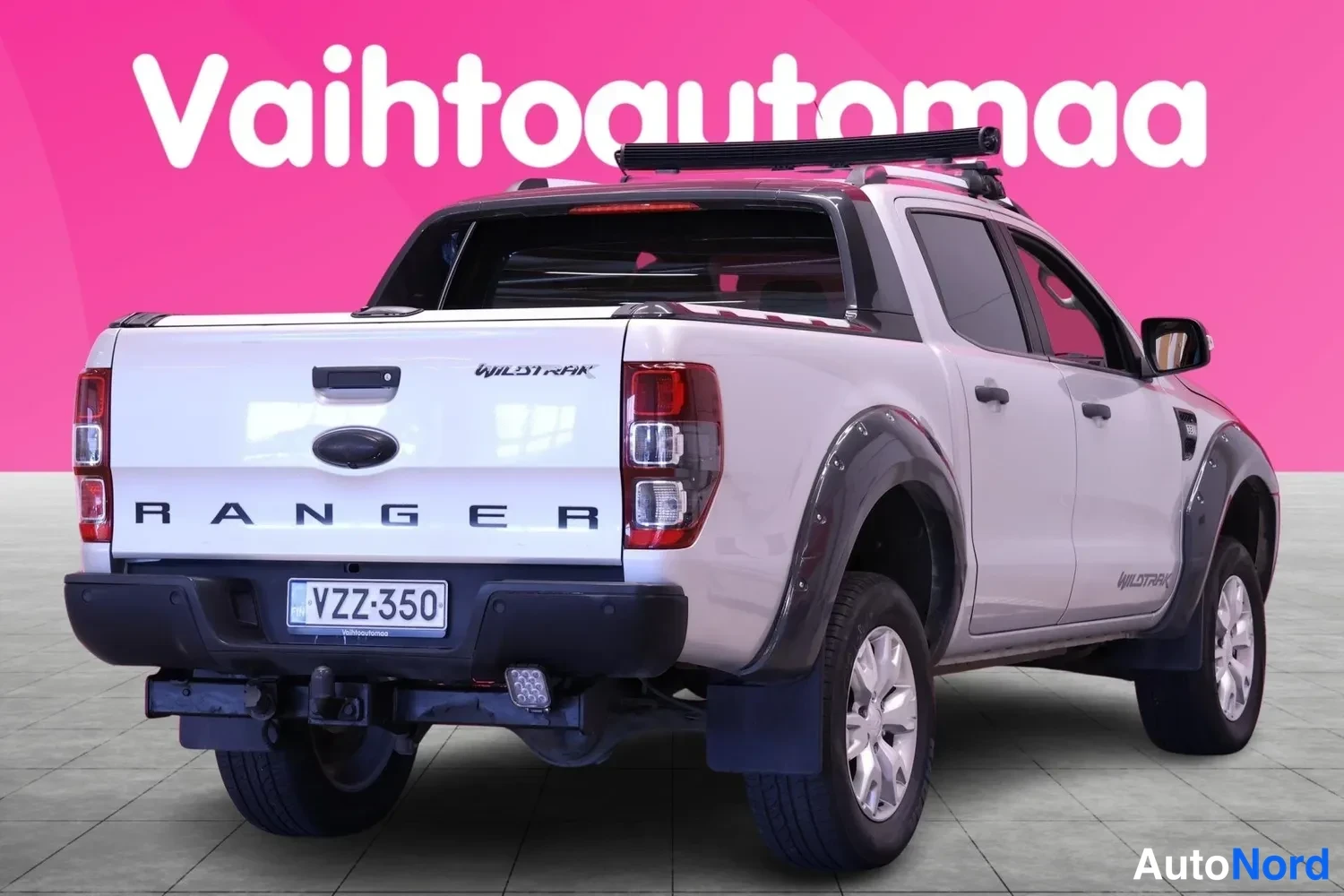 Takaviisto: Valkoisen lavallisen auton takaosan kolmen neljäsosan näkymä; lavan kansilevy kiinni, takapuskassa vakkavalot, taustalla pinkki Vaihtoautomaa-tausta.