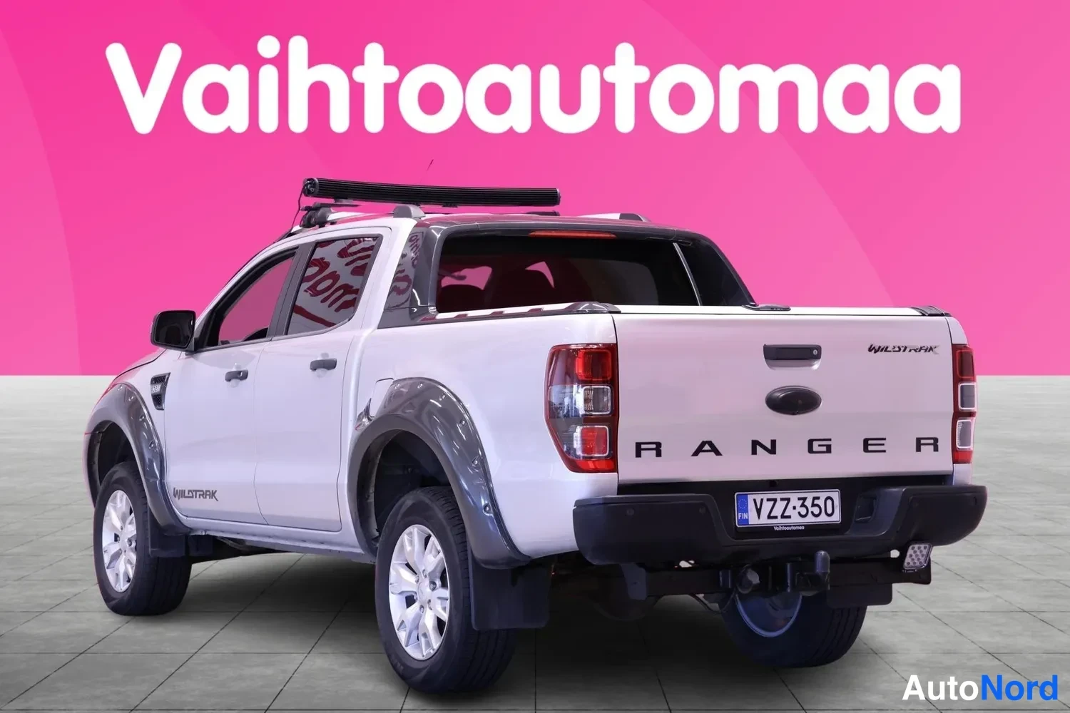 Takaviisto: Ford Ranger -lainen avolava-auto takaa viistosti oikealta kuvattuna, vaaleanpunaisella taustalla. Takapenkin erilliset istuimet näkyvät takalasin läpi.