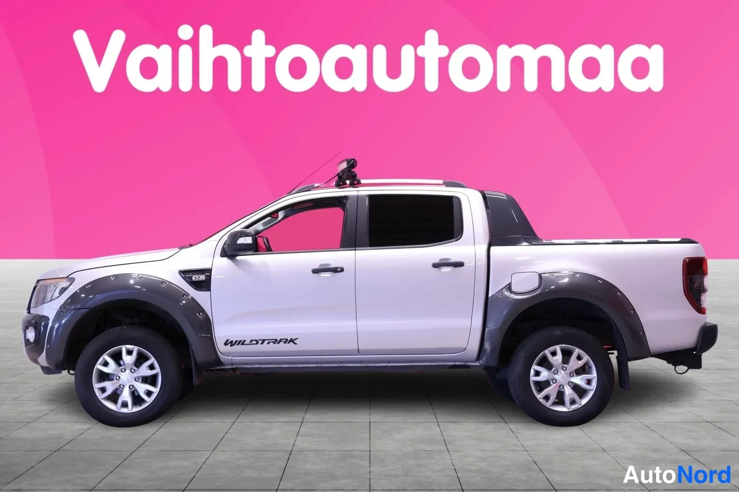 Sivulta: Valkoinen Ford Ranger -tyylinen avolava-auto sivulta vasemmasta suunnasta kuvattuna, vaaleanpunainen tausta. Kuvassa näkyy neljä ovea ja tummat levykkeet.