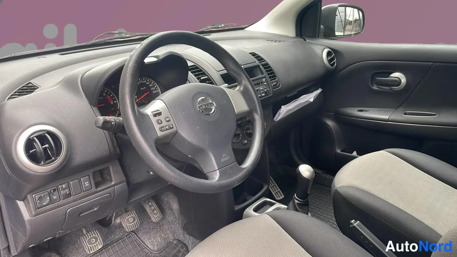 Ohjaamo: Interiørkuvva av førersetet, ratt med Nissan-logo, manuell girspak og dashbord på sort grå farge.